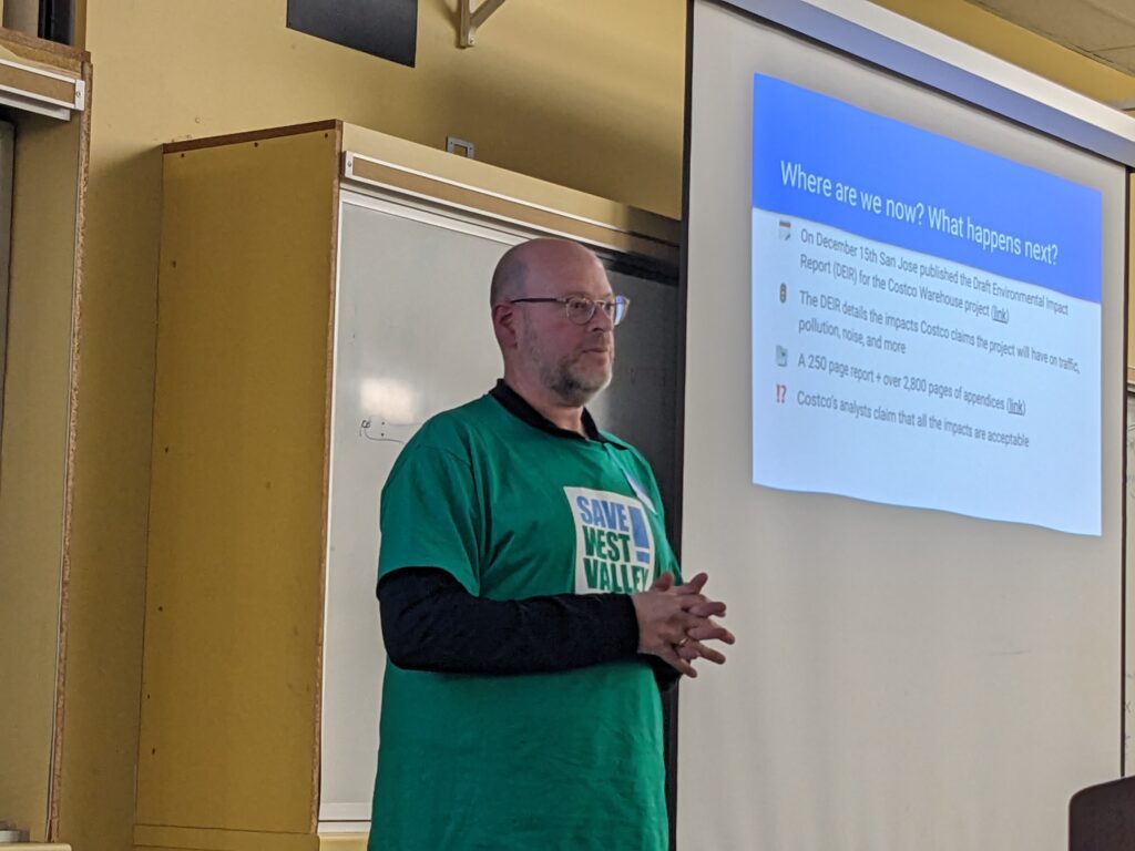 A bald man with a green shirt that reads Save West Valley! and glasses stands at the front of a classroom in front of a slideshow projection. The photo is from his torso up.